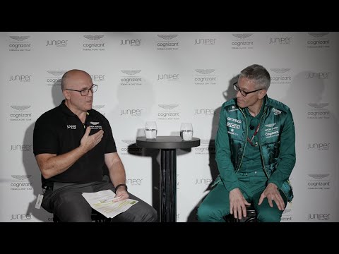 A still shot from the discussion between Juniper’s Darrin Iatrou and Formula One’s Aston Martin team principal, Mike Krack. Darrin is sitting to the left of Mike. They are both sitting on stools with a table between theme. There is a stop-and-repeat banner behind them with the Juniper Networks logo and cognizant logo. Darrin appears to be talking or asking a question. Darrin is wearing a black collared shirt with the Aston Martin and Juniper Networks logos. Mike is wearing a teal jumpsuit. The top is shiny and has the team’s partners in white written on the front.